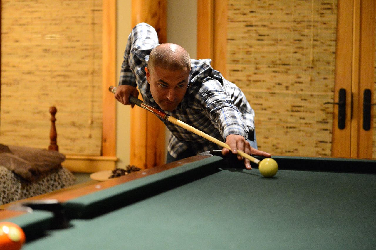 A man in a plaid shirt leans over a pool table, aiming his cue stick at the yellow ball.