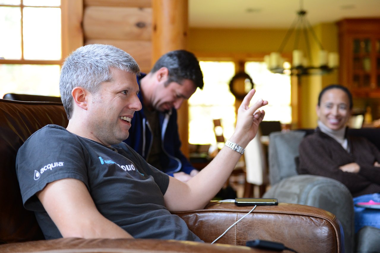A man in an Acquia t-shirt sits on a couch, smiling and gesturing, while others in the background react.