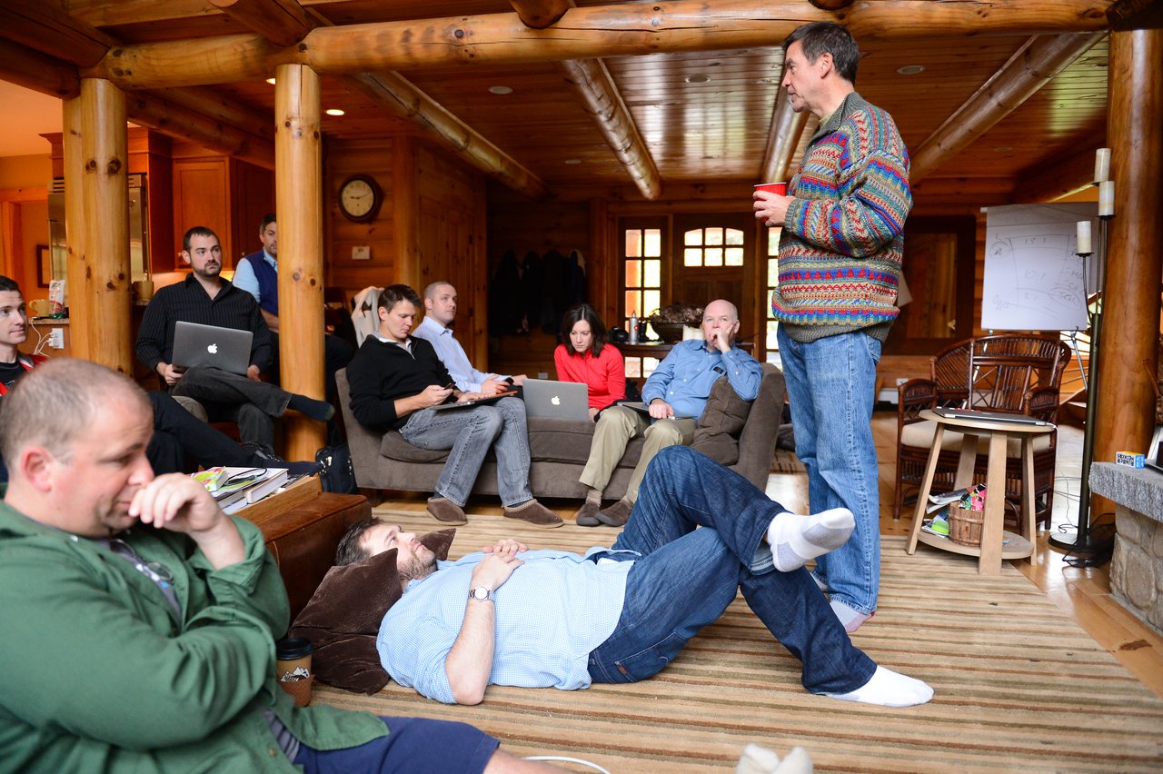 A group of people in a cabin, some using laptops, while one person stands and speaks holding a red cup.