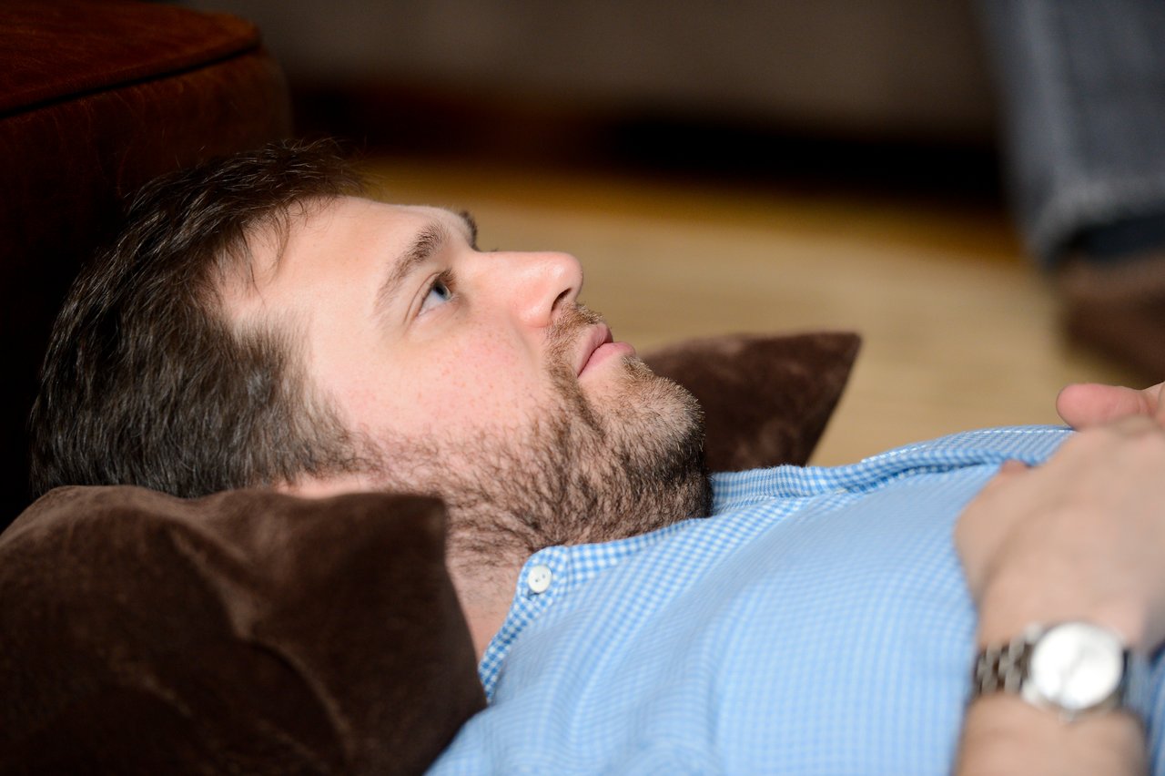 A man in a blue shirt lies on his back with a pillow under his head, looking upward.
