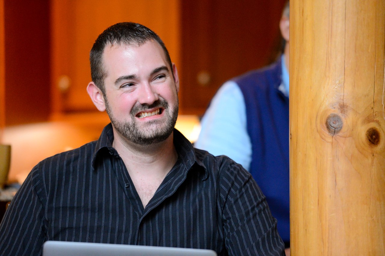 A man in a black shirt sits at a laptop, smiling and looking to the side.