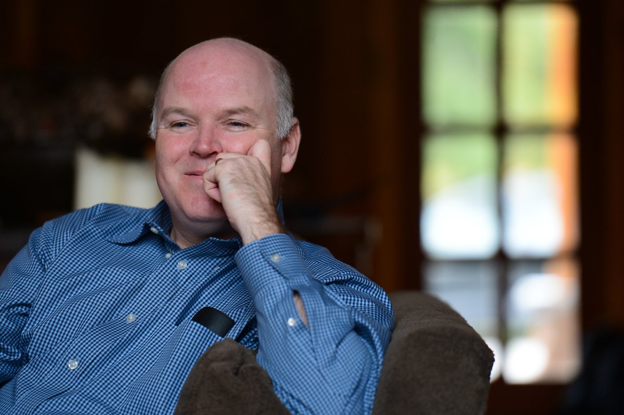 A man in a blue shirt sits on a couch, resting his chin on his hand and smiling.