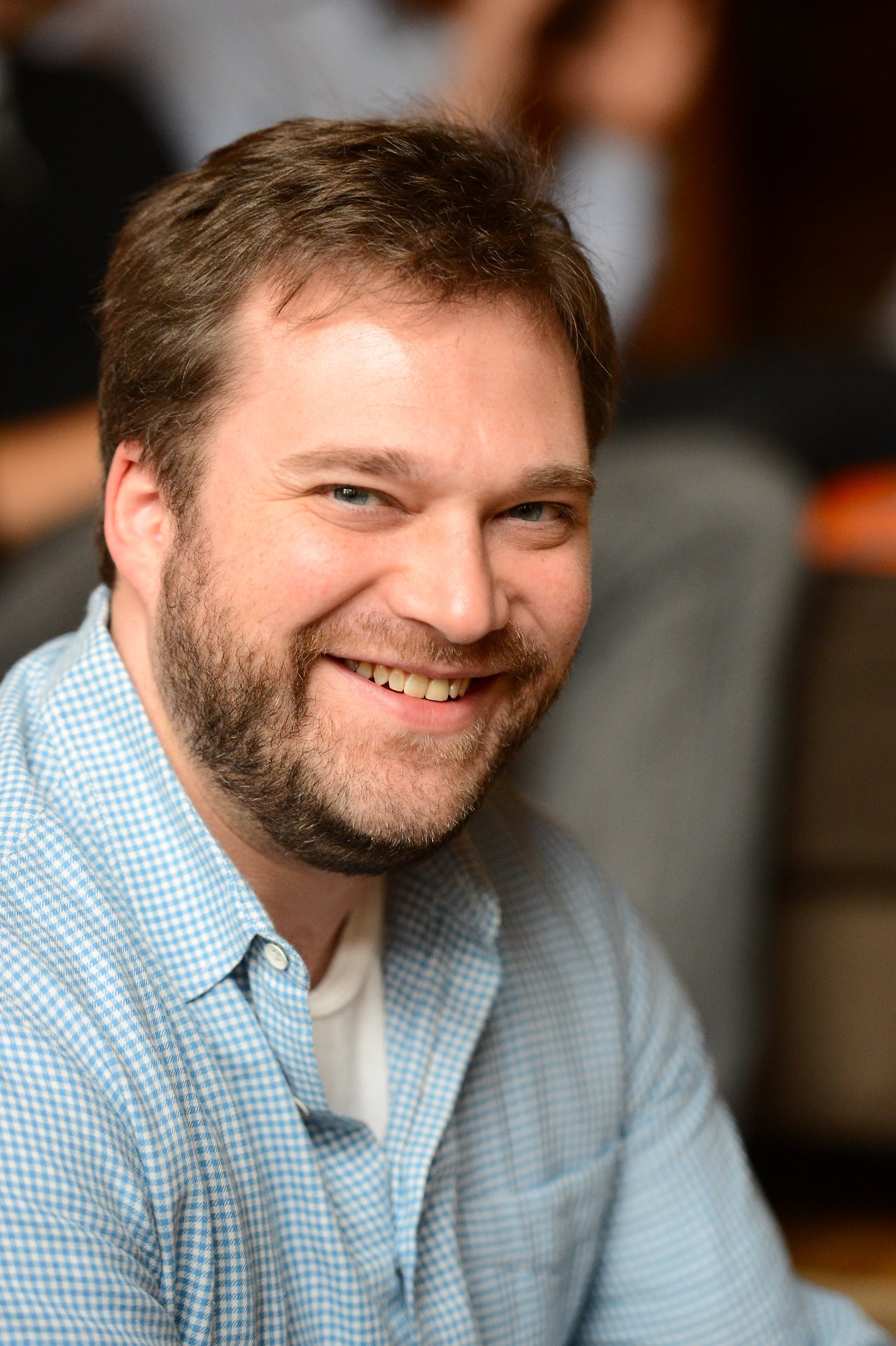 A man with a beard and a blue checkered shirt smiles at the camera in a casual setting.