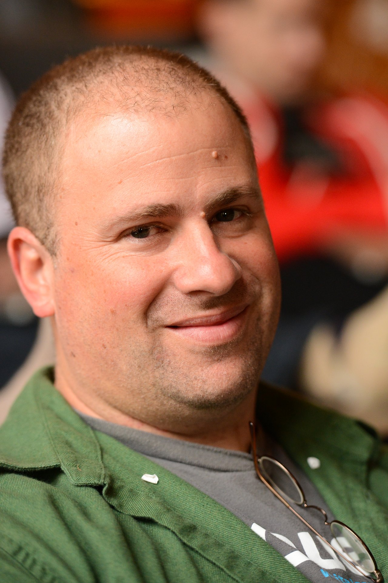 A man in a green jacket smiles at the camera, with glasses hanging from his shirt.