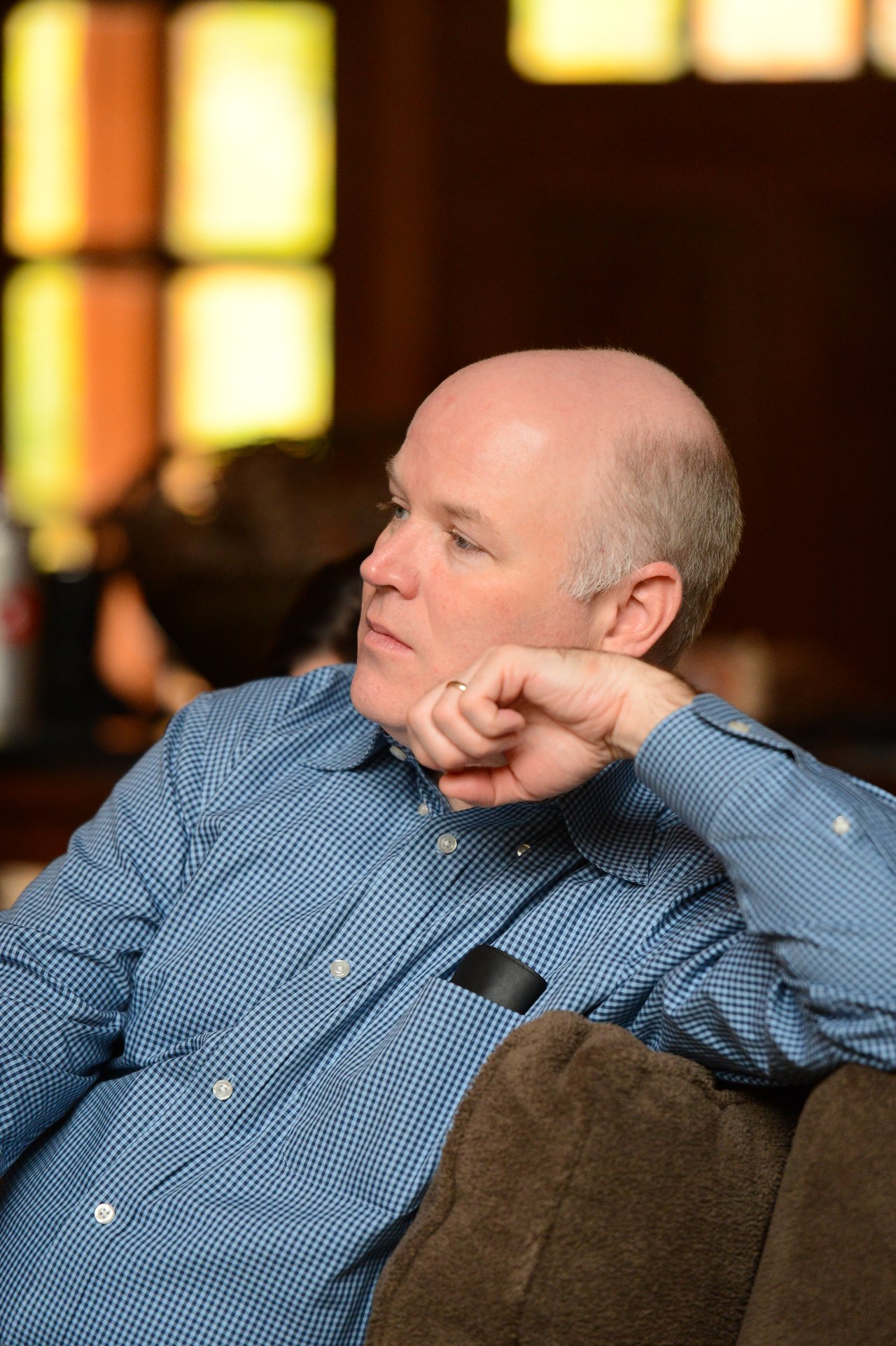 A man in a blue checkered shirt sits on a couch, resting his chin on his hand, looking thoughtful.