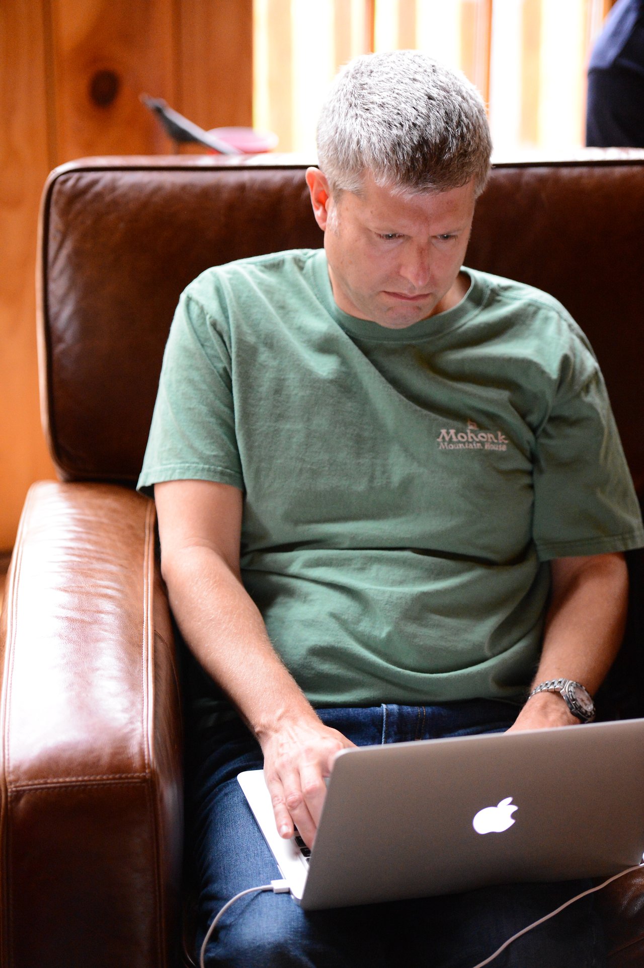 A man in a green shirt sits on a brown leather chair, focused on his laptop.