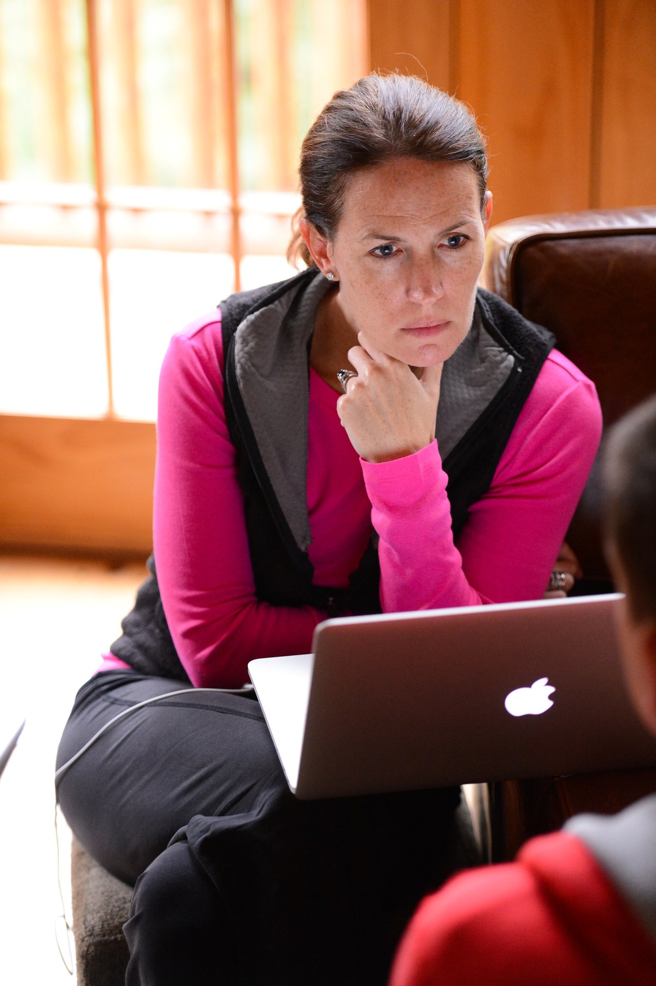A woman in a pink sweater sits with a laptop, listening attentively to someone off-camera.
