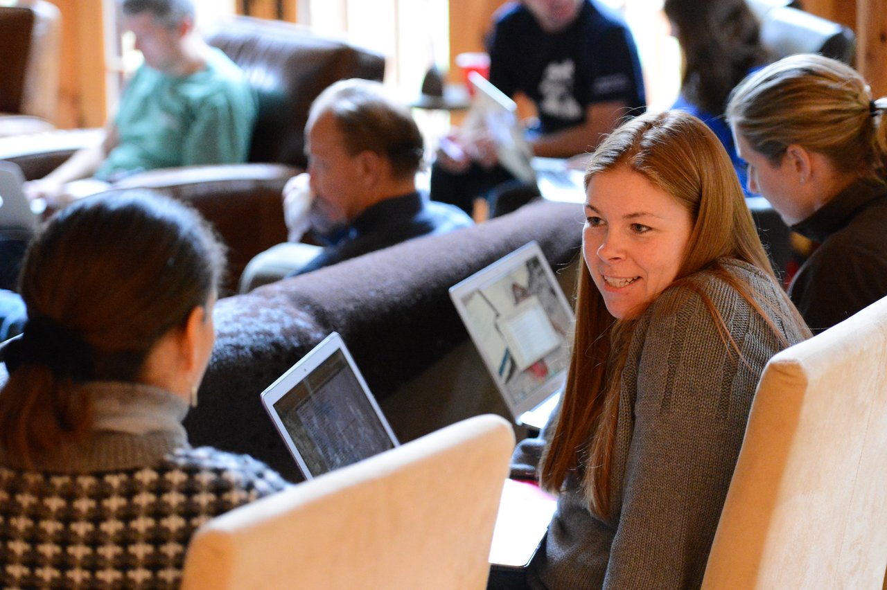 A woman in a gray sweater smiles while talking to a colleague, with laptops open in a group setting.