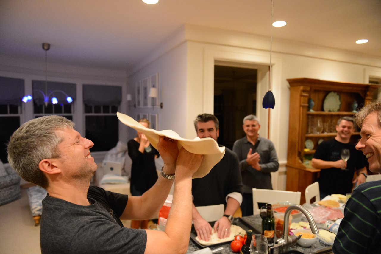 A man smiles while tossing pizza dough in the air as others watch and clap in a kitchen.