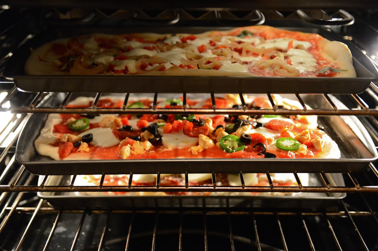 Two homemade pizzas with various toppings bake on metal trays inside an oven.