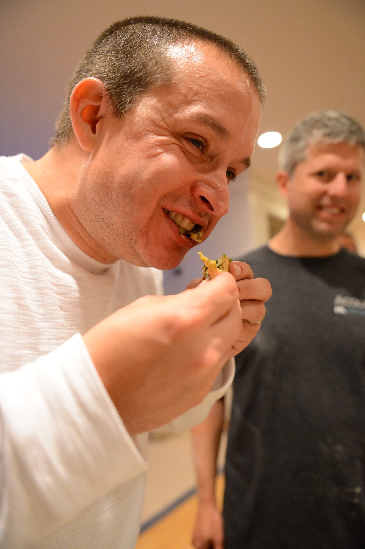 A man in a white shirt takes a bite of food while another person smiles in the background.