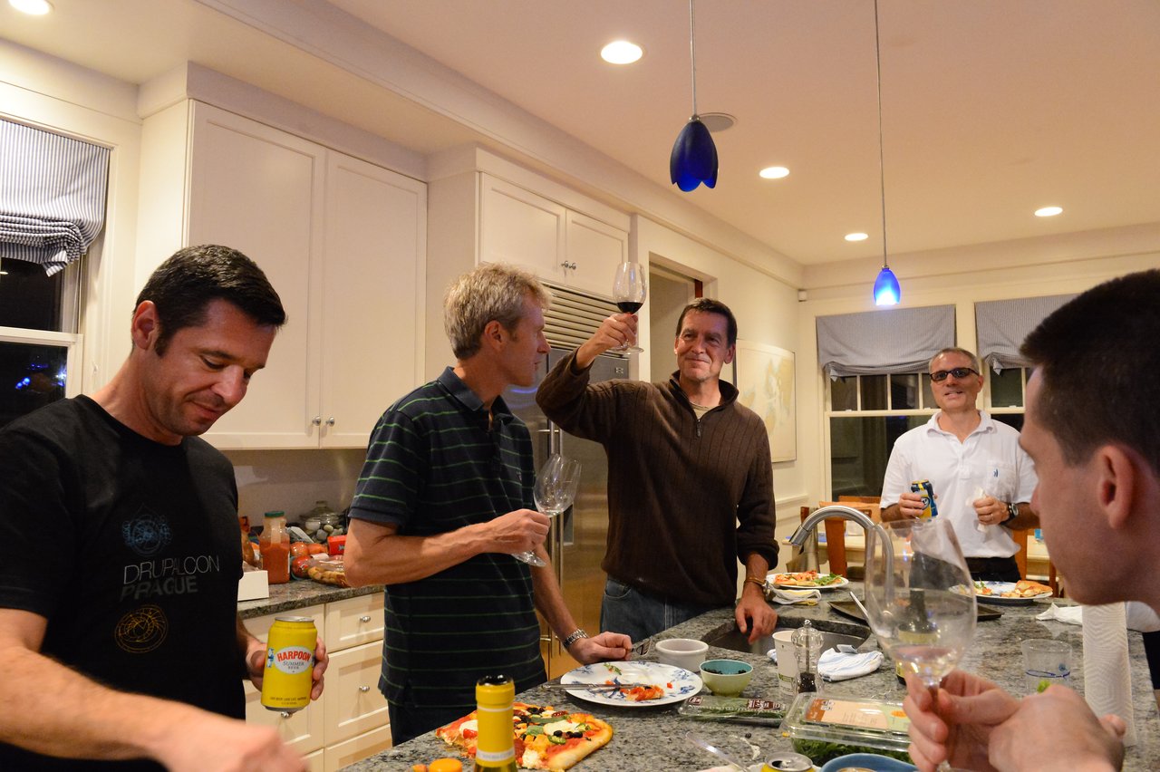 A group of people in a kitchen enjoying food and drinks, with one person raising a glass in a toast.
