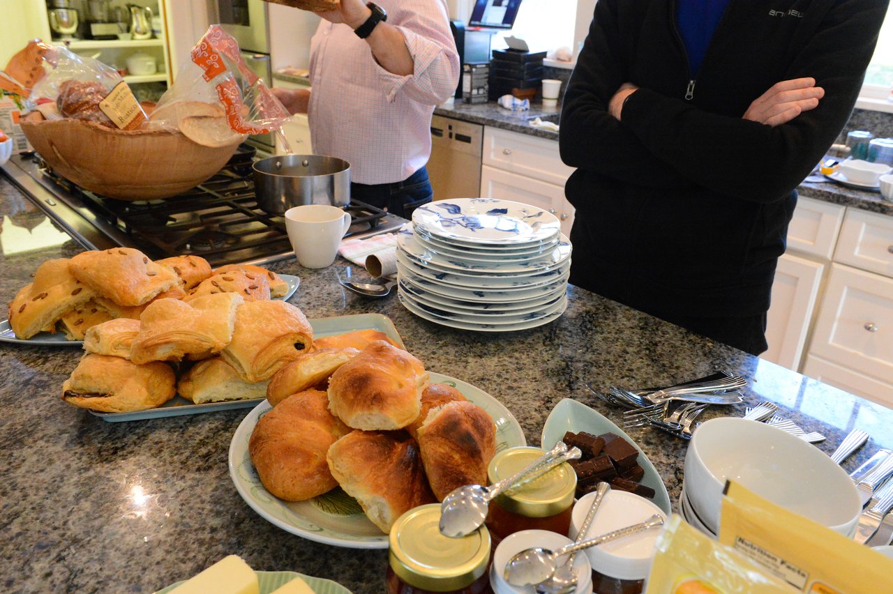 A kitchen counter with plates, pastries, jam jars, and utensils, while two people stand nearby.