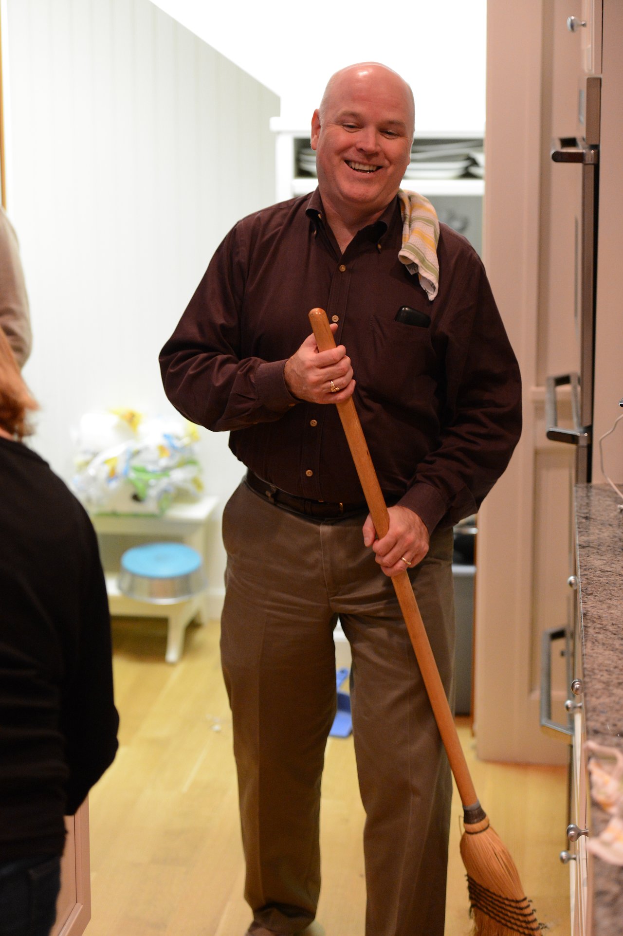 A smiling man in a dark shirt holds a broom in a kitchen, with a towel over his shoulder.