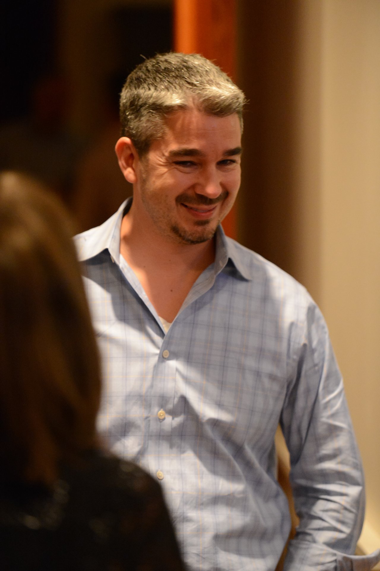A man in a light blue shirt smiles while talking to someone at an indoor event.