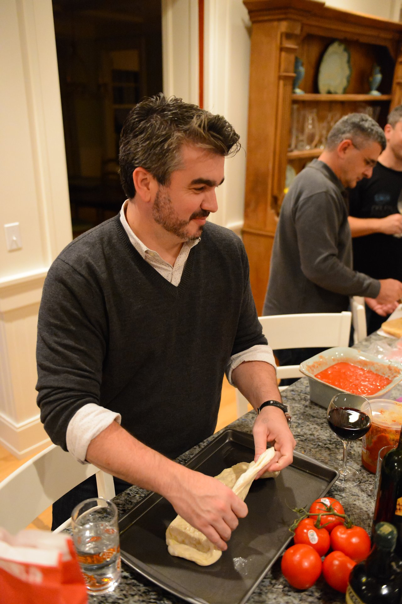 A man stretches dough on a baking tray while preparing food in a kitchen with others.