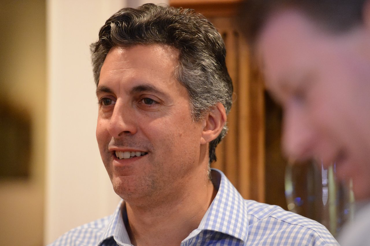 A man in a checkered shirt smiles while another person in the foreground is slightly out of focus.