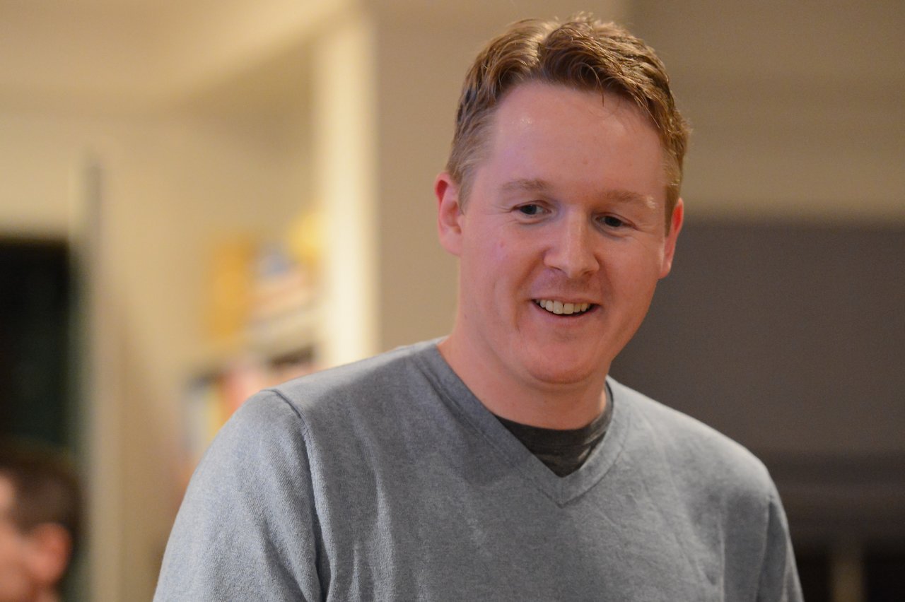 A man in a gray sweater smiles while looking downward in an indoor setting.