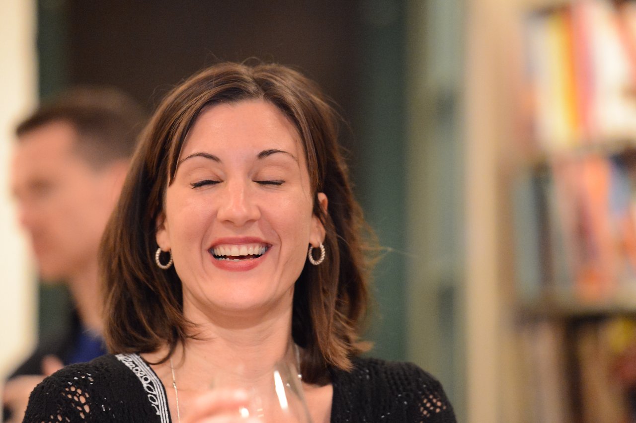 A woman with shoulder-length brown hair laughs with her eyes closed, holding a glass in her hand.