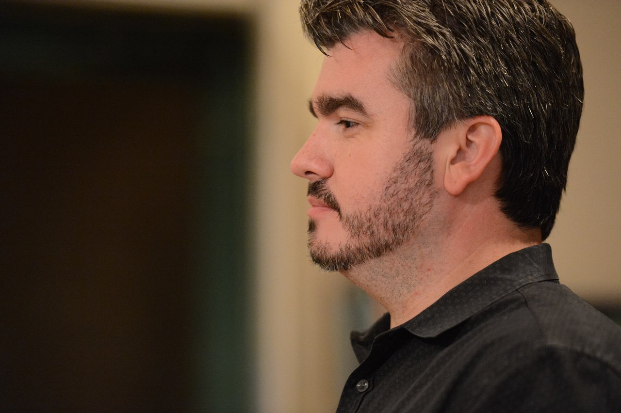 A man in a black shirt looks attentively to the side in an indoor setting.