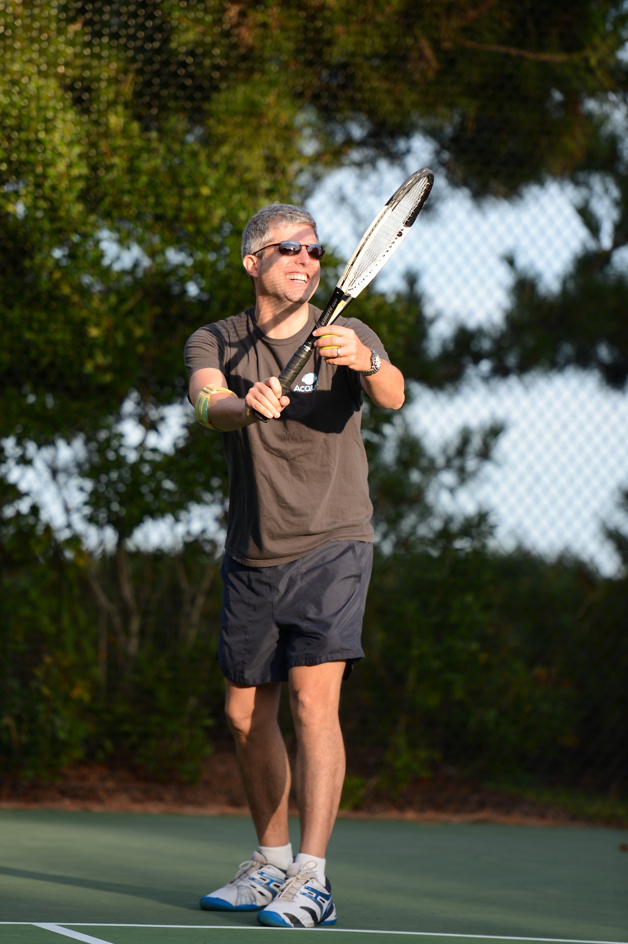 A man wearing sunglasses and a t-shirt plays tennis, holding a racket and a ball on an outdoor court.