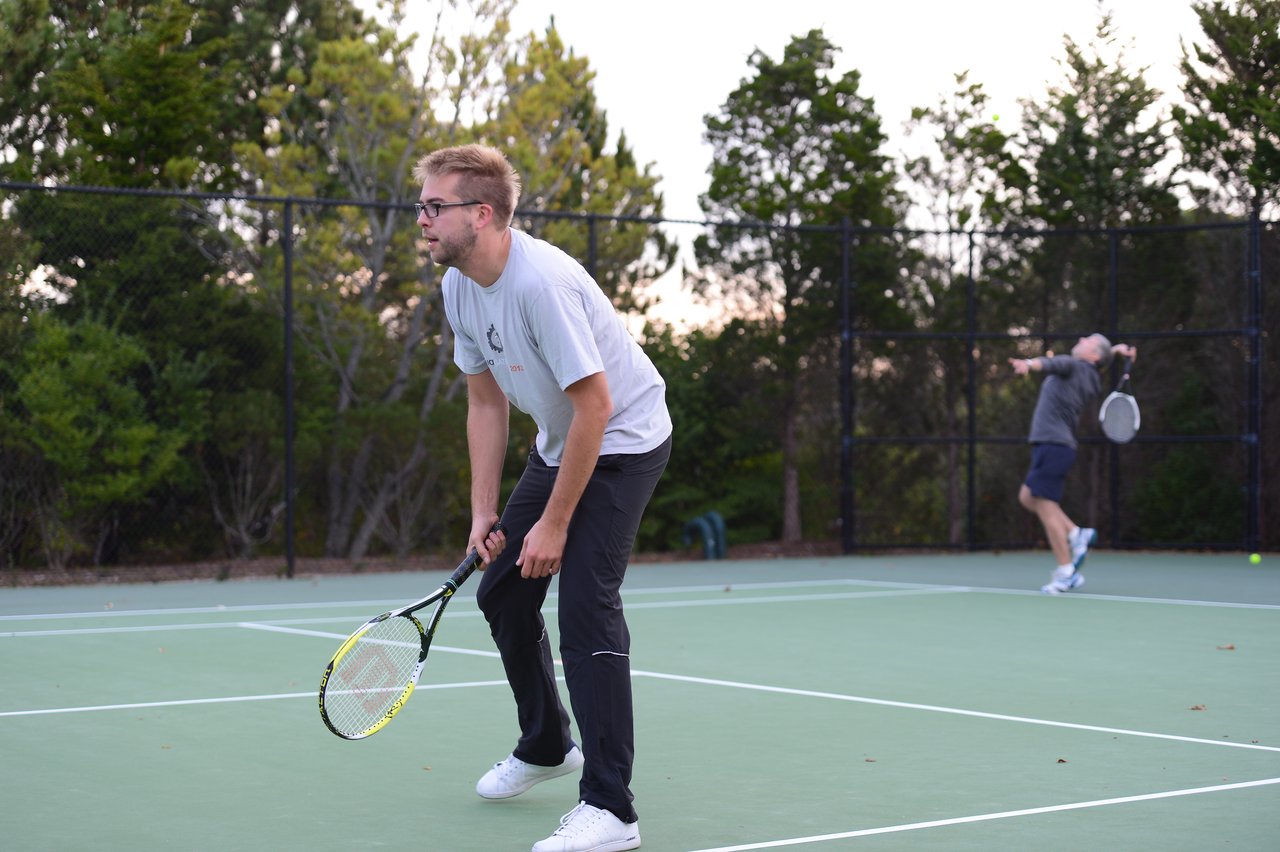 Two people playing tennis on an outdoor court, one preparing to receive a serve while the other hits the ball.