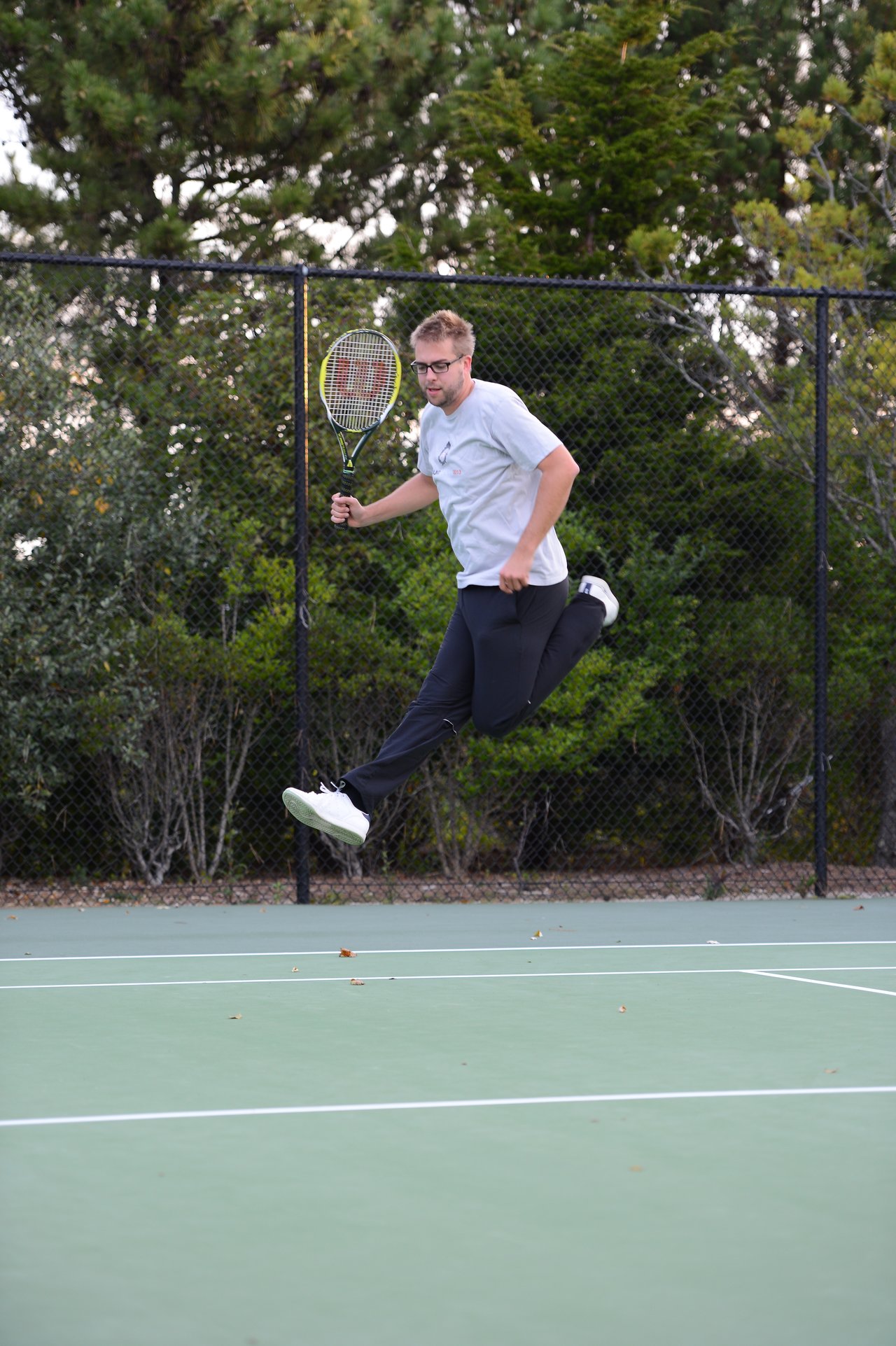 A man in glasses and a gray shirt jumps while holding a tennis racket on an outdoor court.