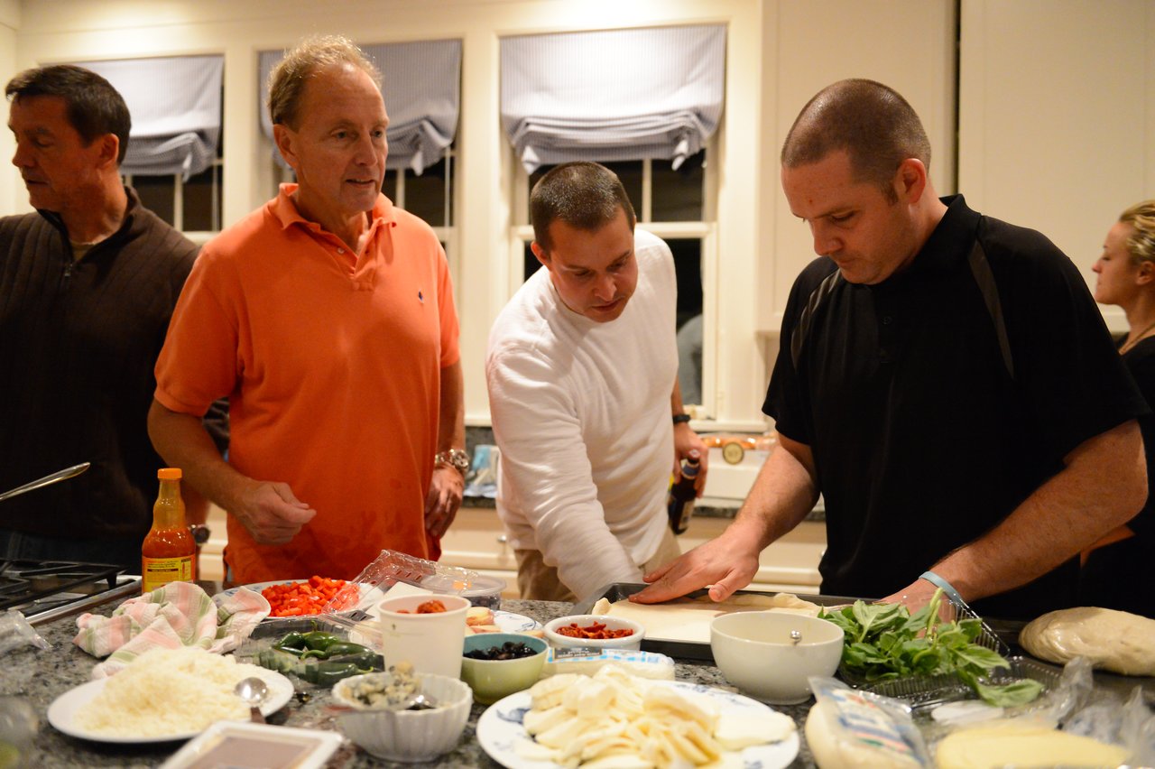 A group of people prepares food in a kitchen, with one person rolling dough while others assist and observe.
