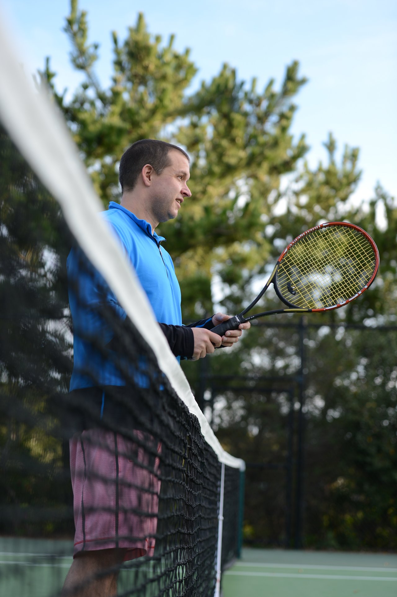 A man in a blue shirt holds a tennis racket while standing near the net on an outdoor court.