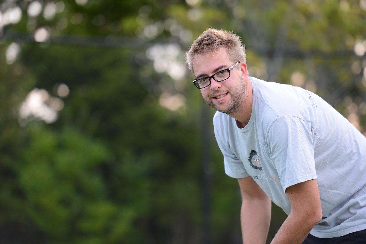 A man wearing glasses and a light-colored t-shirt leans forward and smiles outdoors with trees in the background.