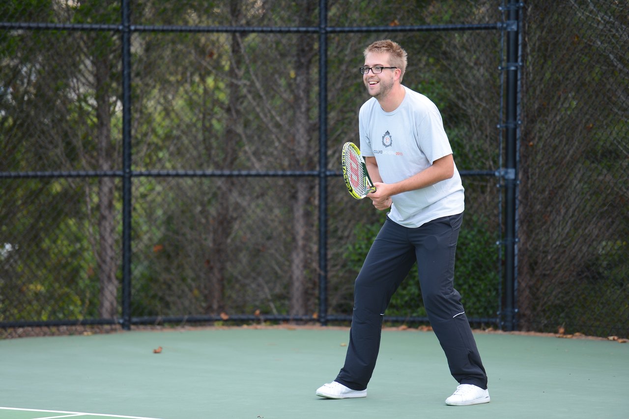A man in a white t-shirt and black pants holds a tennis racket, preparing to play on an outdoor court.