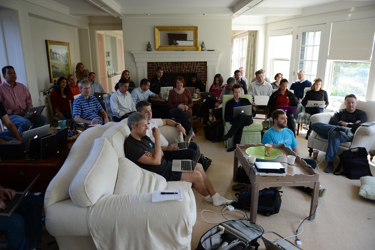 A group of people in a living room, using laptops and listening to a presentation at an Acquia offsite event.