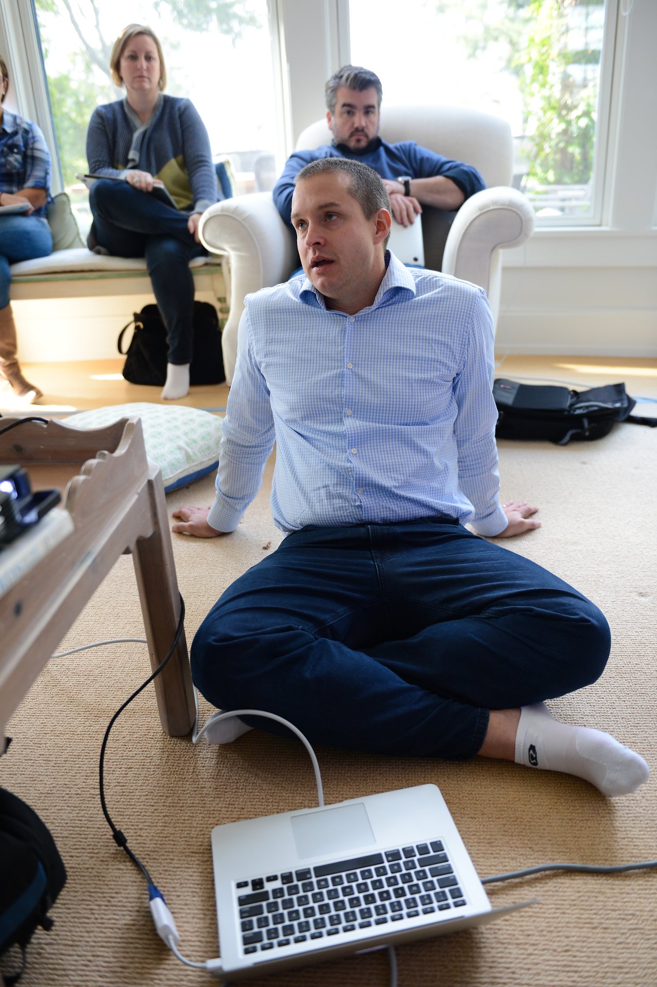 A man in a blue shirt sits on the floor, speaking, with a laptop and cables nearby.