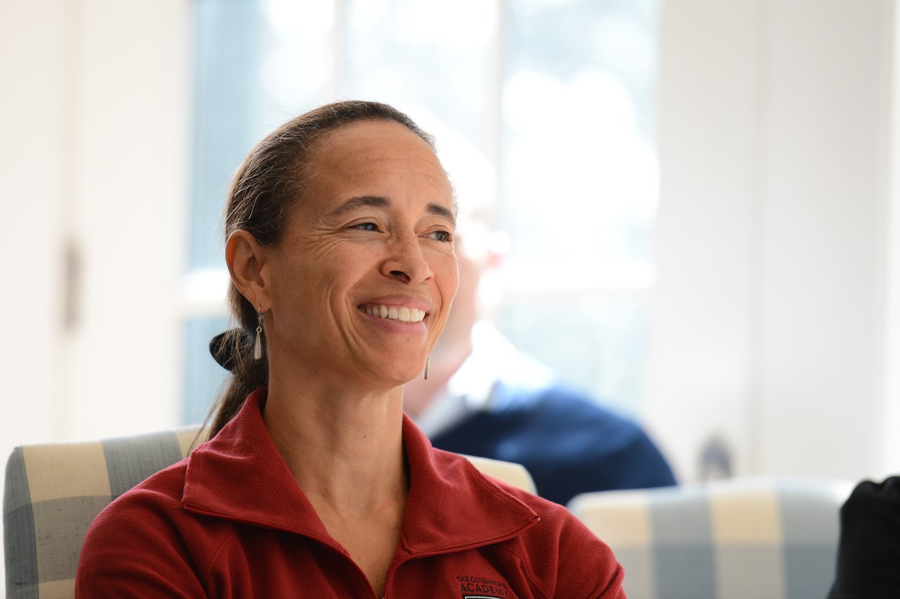 A woman in a red jacket smiles while sitting indoors, with a blurred person in the background.