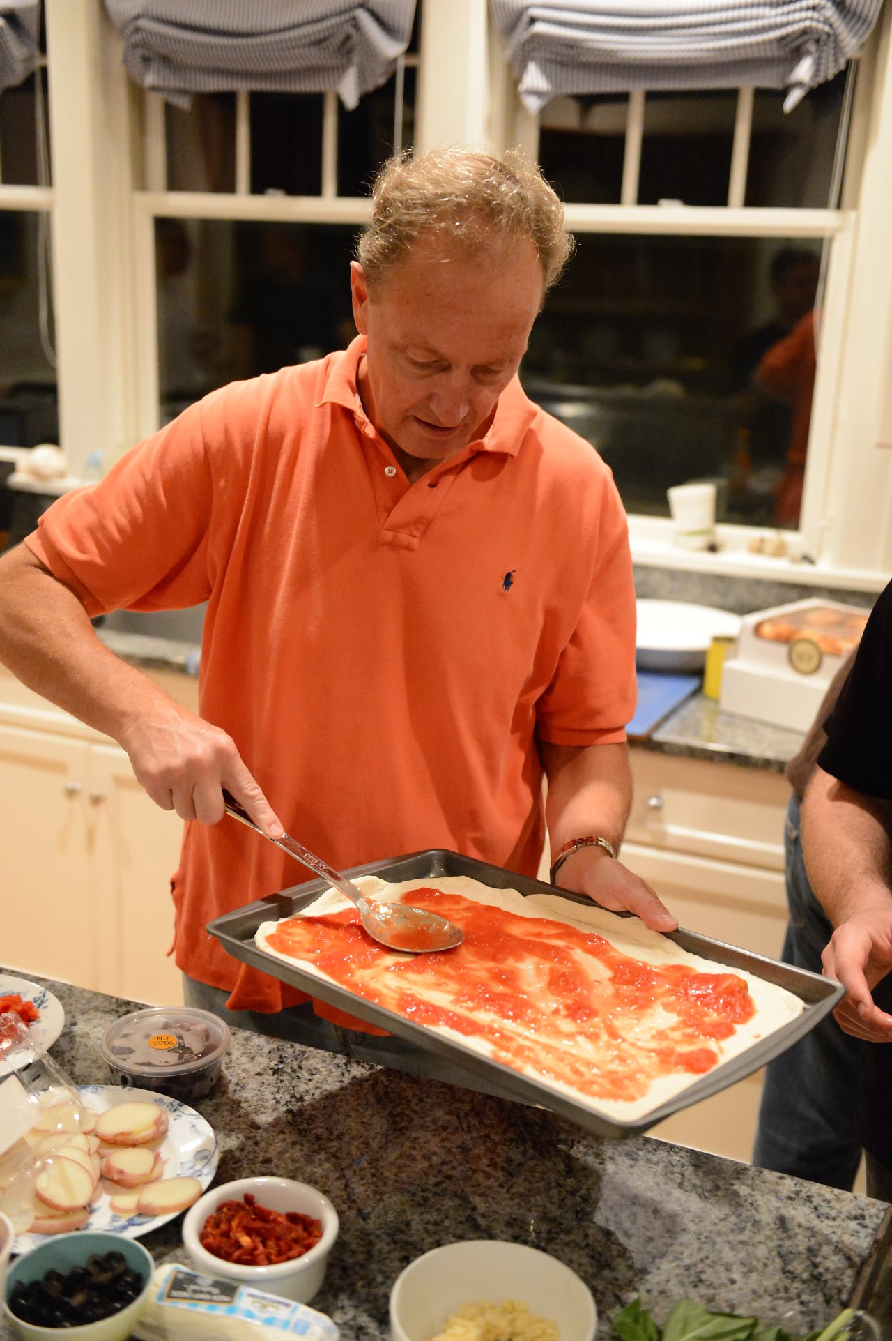 A man in an orange shirt spreads tomato sauce on pizza dough in a kitchen.