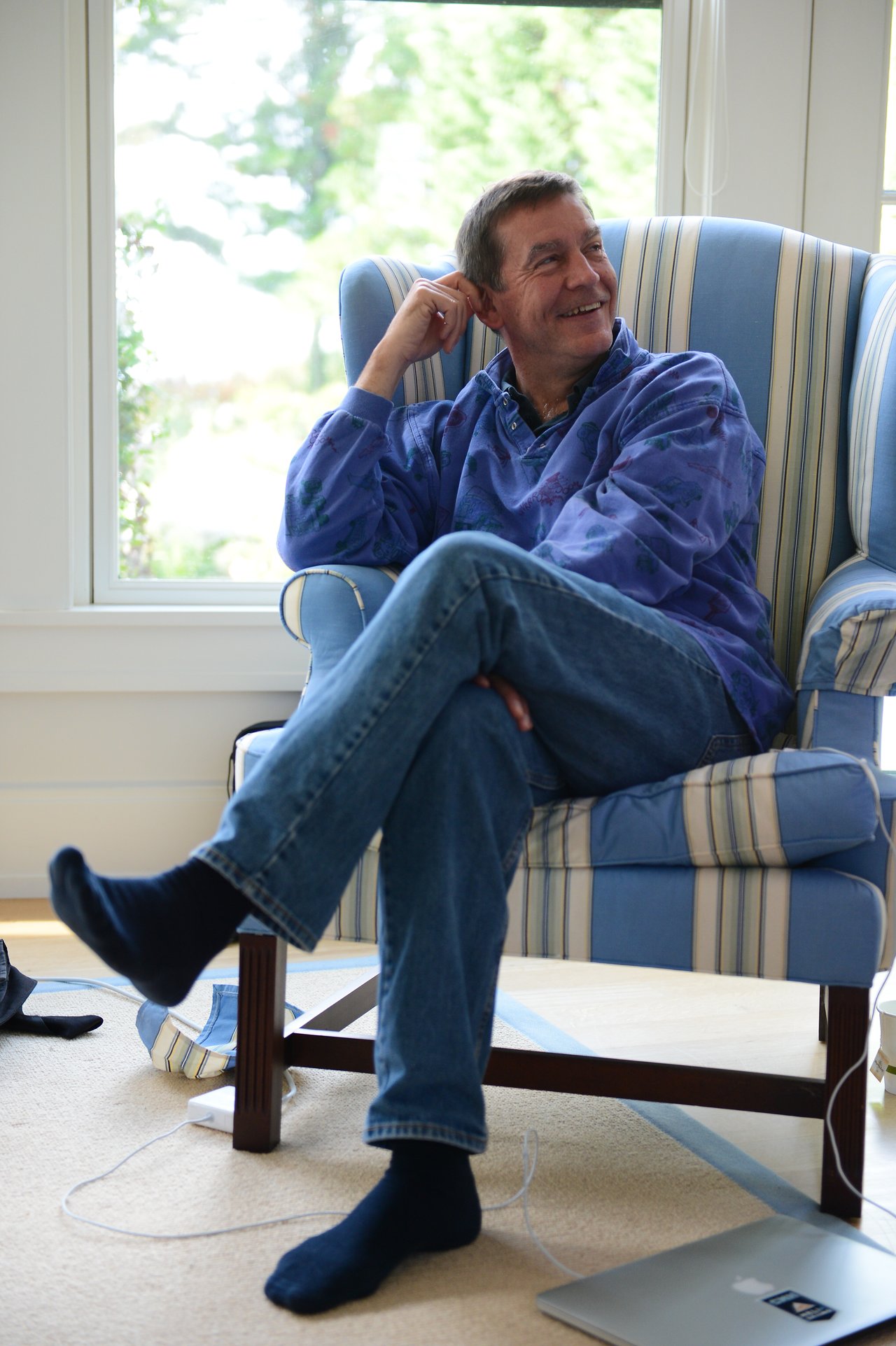 A man in a blue sweater sits in a striped armchair, smiling and listening with a relaxed posture.