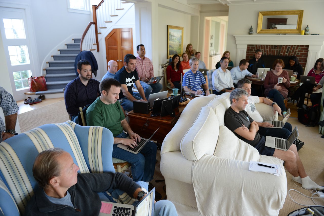 A group of people in a living room setting, working on laptops and listening to a presentation or discussion.
