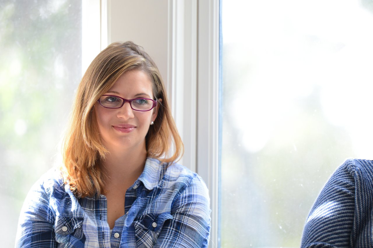 A woman wearing glasses and a plaid shirt sits near a window, smiling slightly.