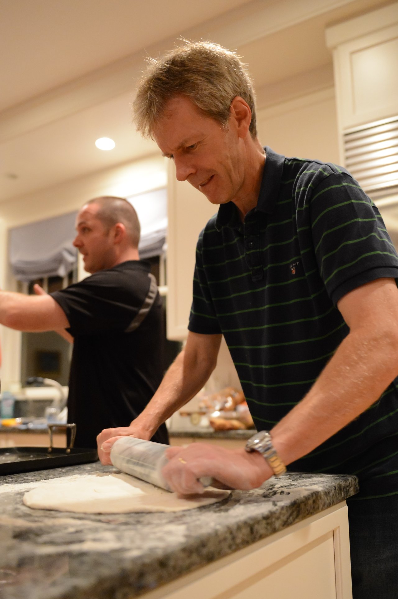 A man in a striped shirt rolls out dough on a kitchen counter, while another person gestures behind him.