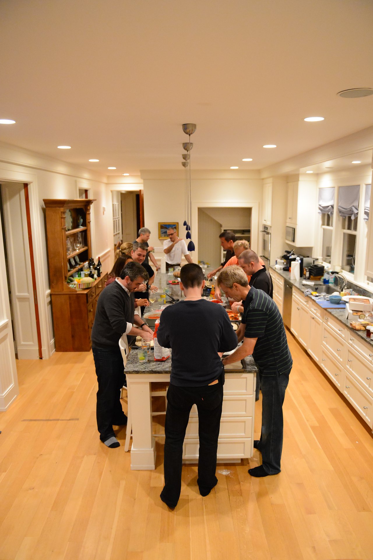 A group of people gathers around a kitchen island, preparing food together in a well-lit home setting.