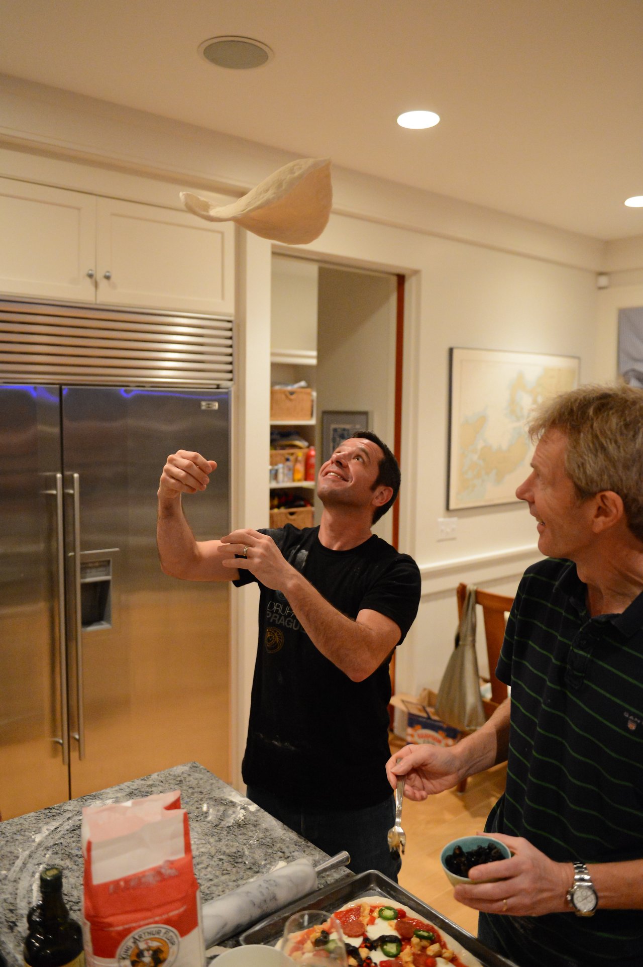 Two people making pizza in a kitchen, one tossing dough in the air while the other adds toppings.