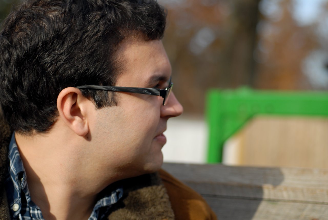 A man wearing glasses and a brown jacket looks to the side while sitting on a wooden bench outdoors.
