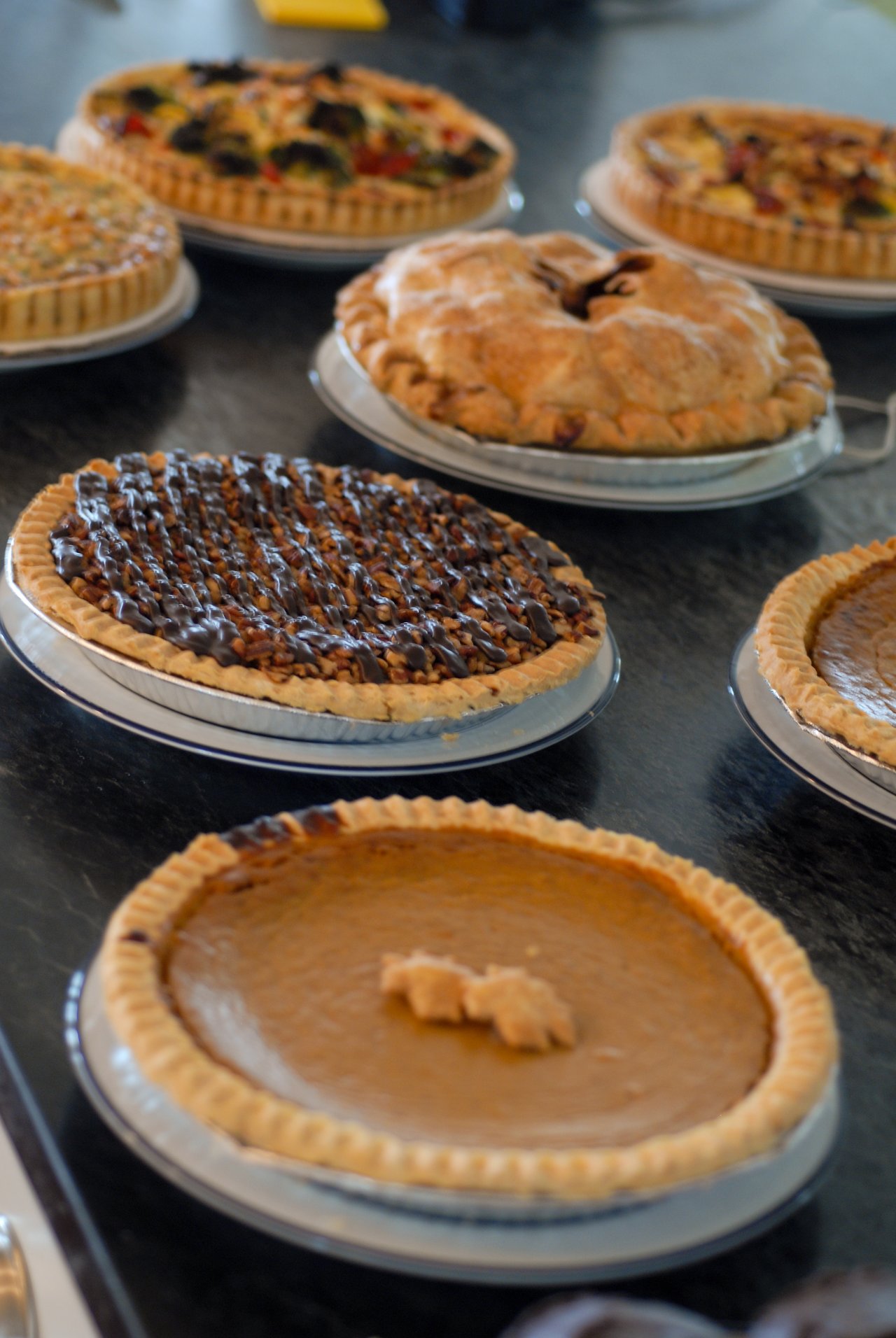 Several cakes and tarts, including chocolate-drizzled and pumpkin pies, are displayed on a table in metal trays.