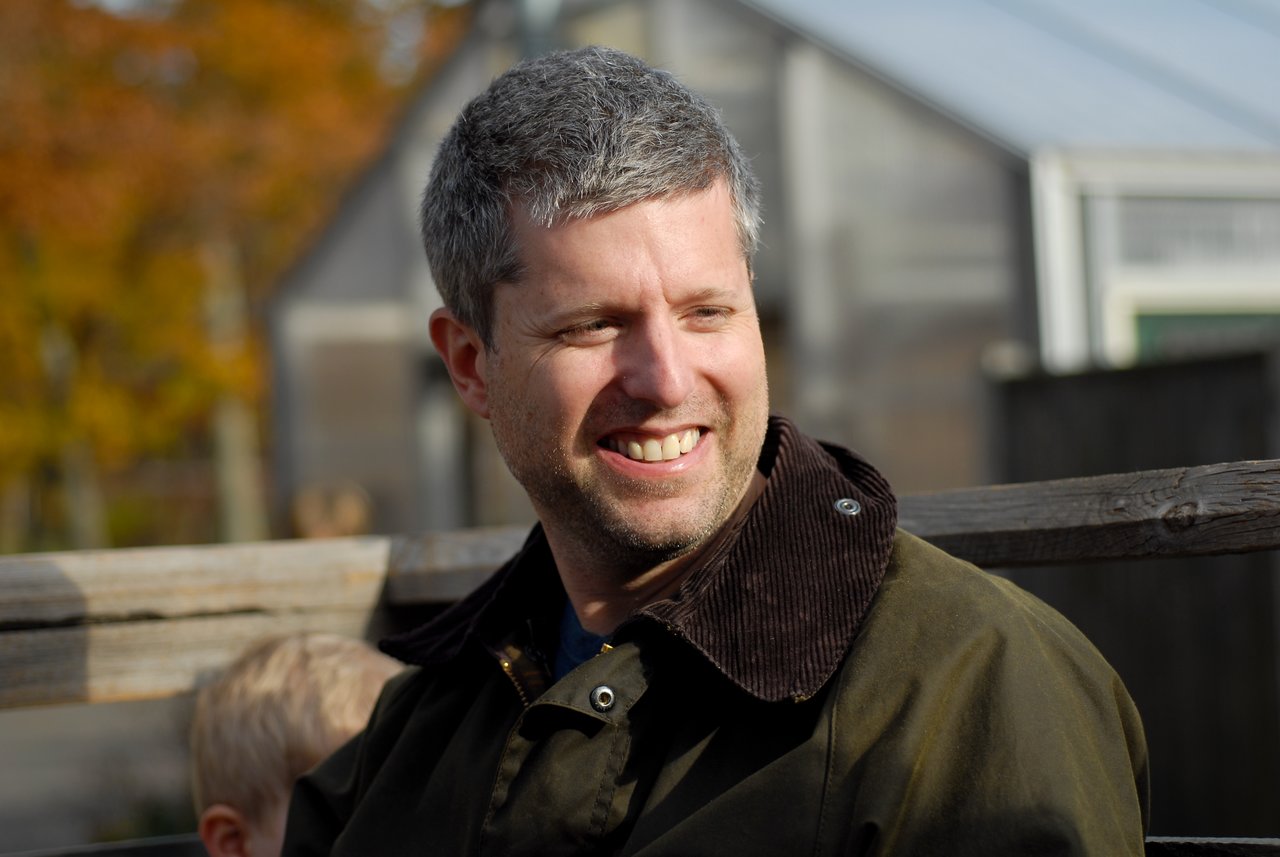 A man with short gray hair and a green jacket smiles while sitting outdoors on a wooden bench.