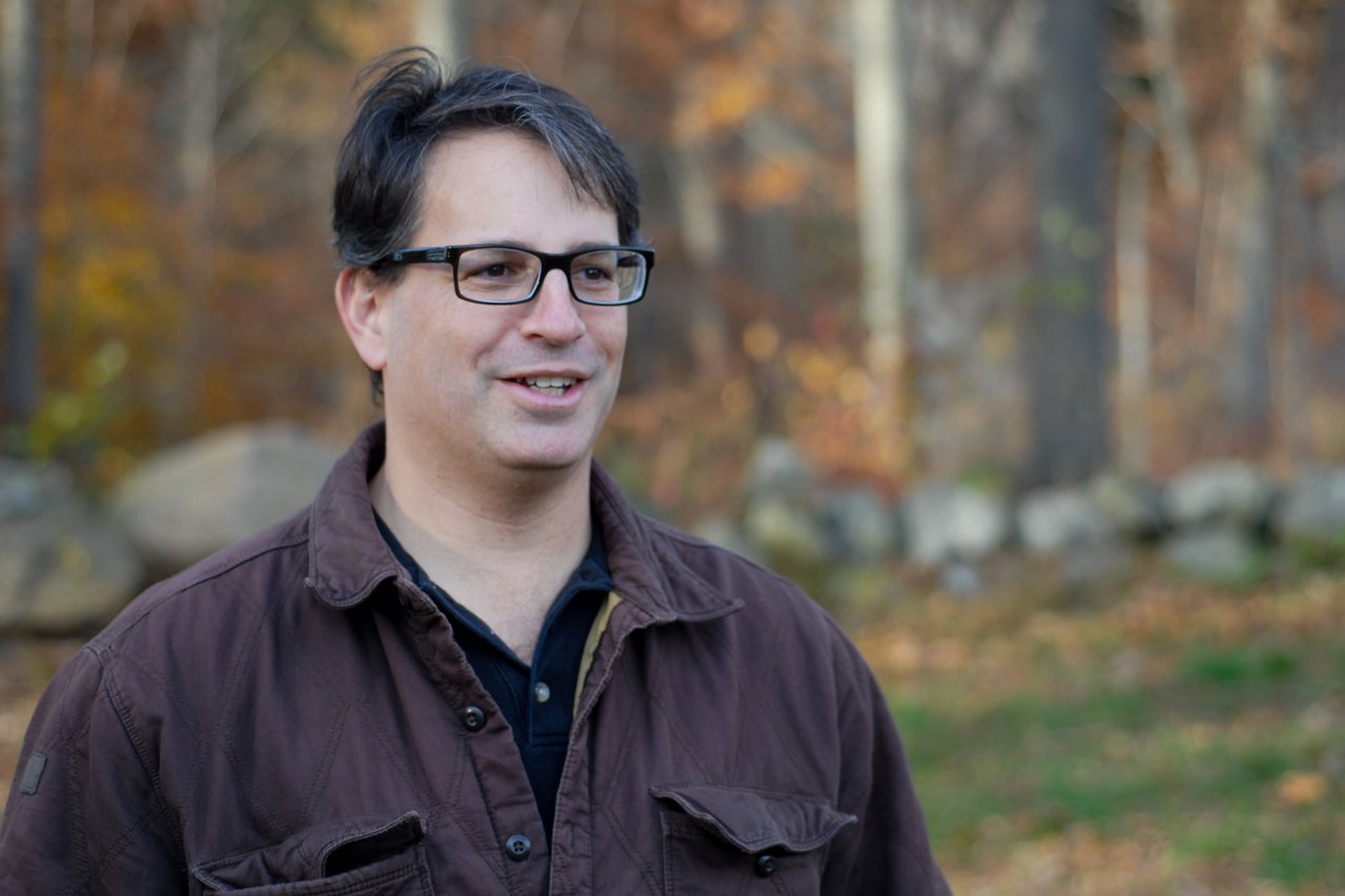 A man wearing glasses and a brown jacket smiles while standing outdoors in a wooded area with autumn colors.