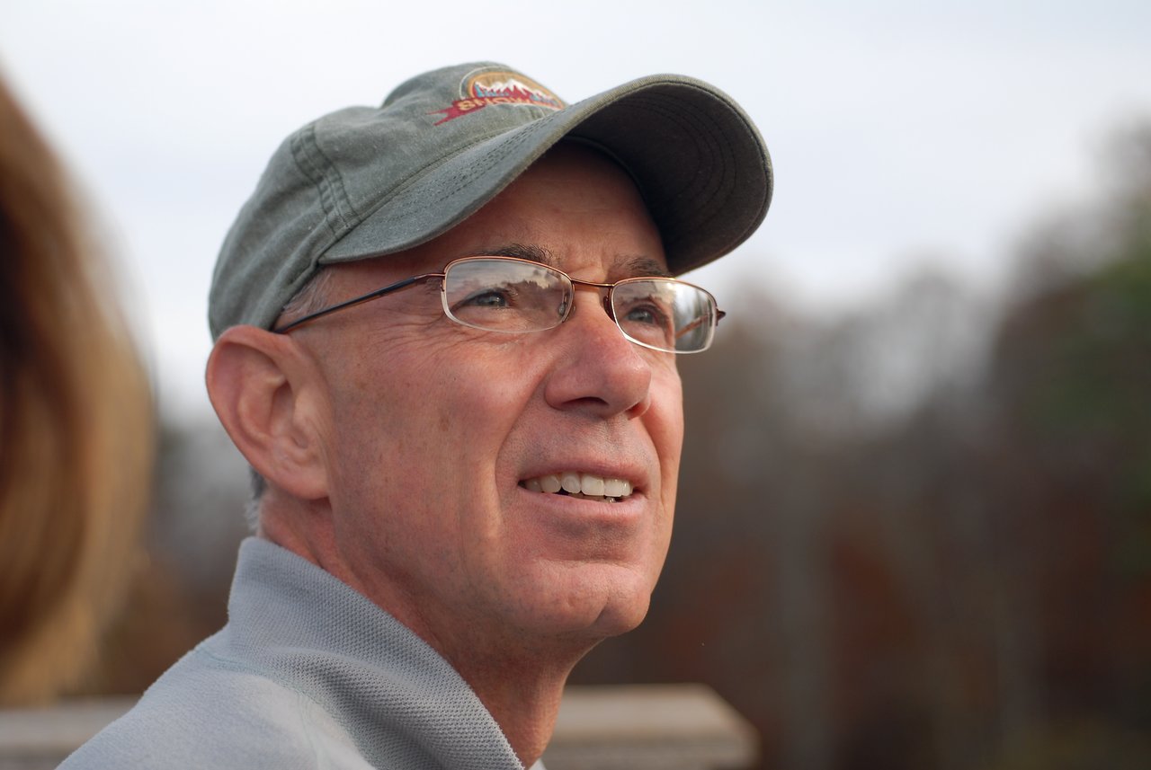 A man wearing glasses and a cap looks off into the distance with a slight smile.