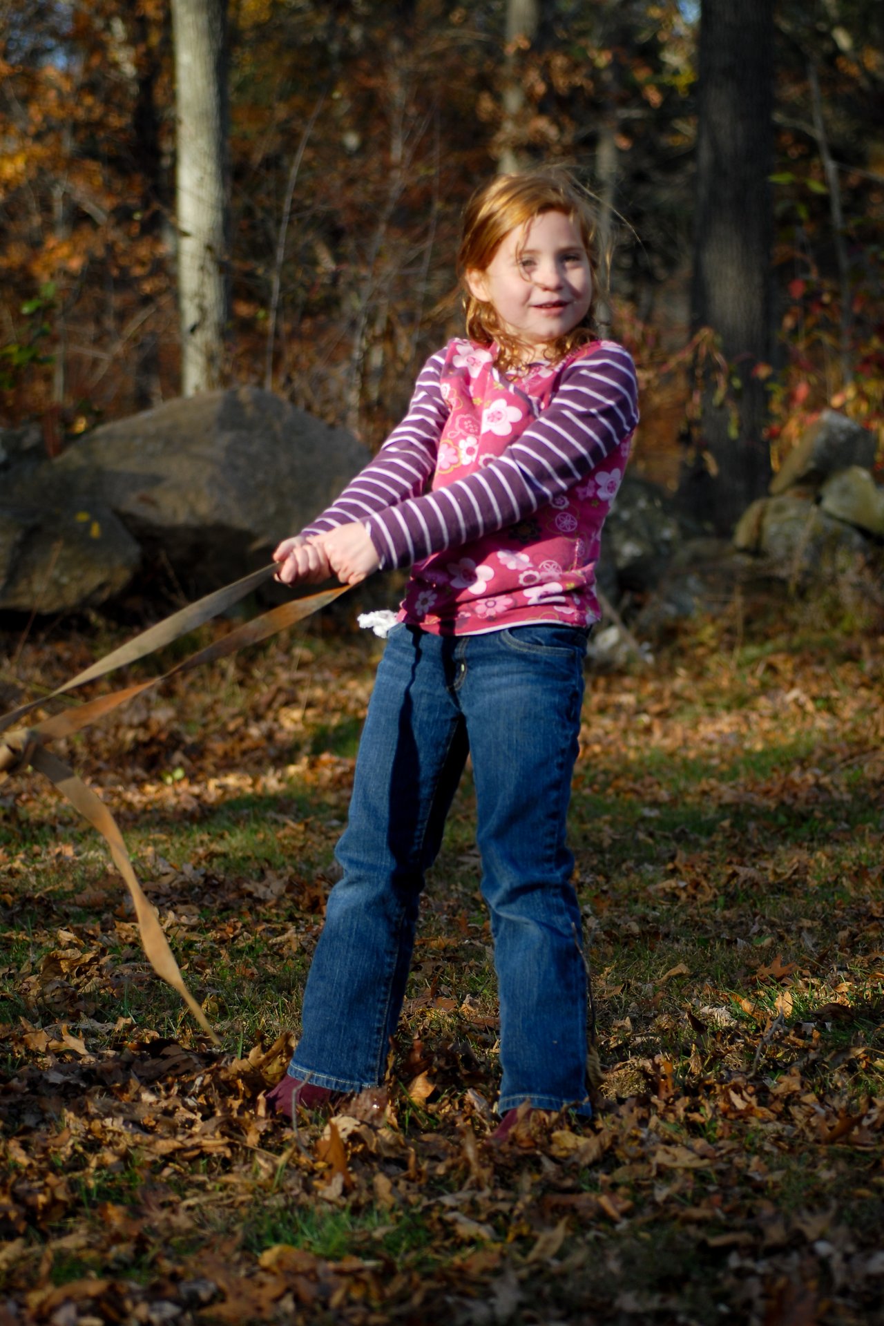 A young girl in a pink and purple outfit pulls on a long ribbon outdoors in an autumn setting.