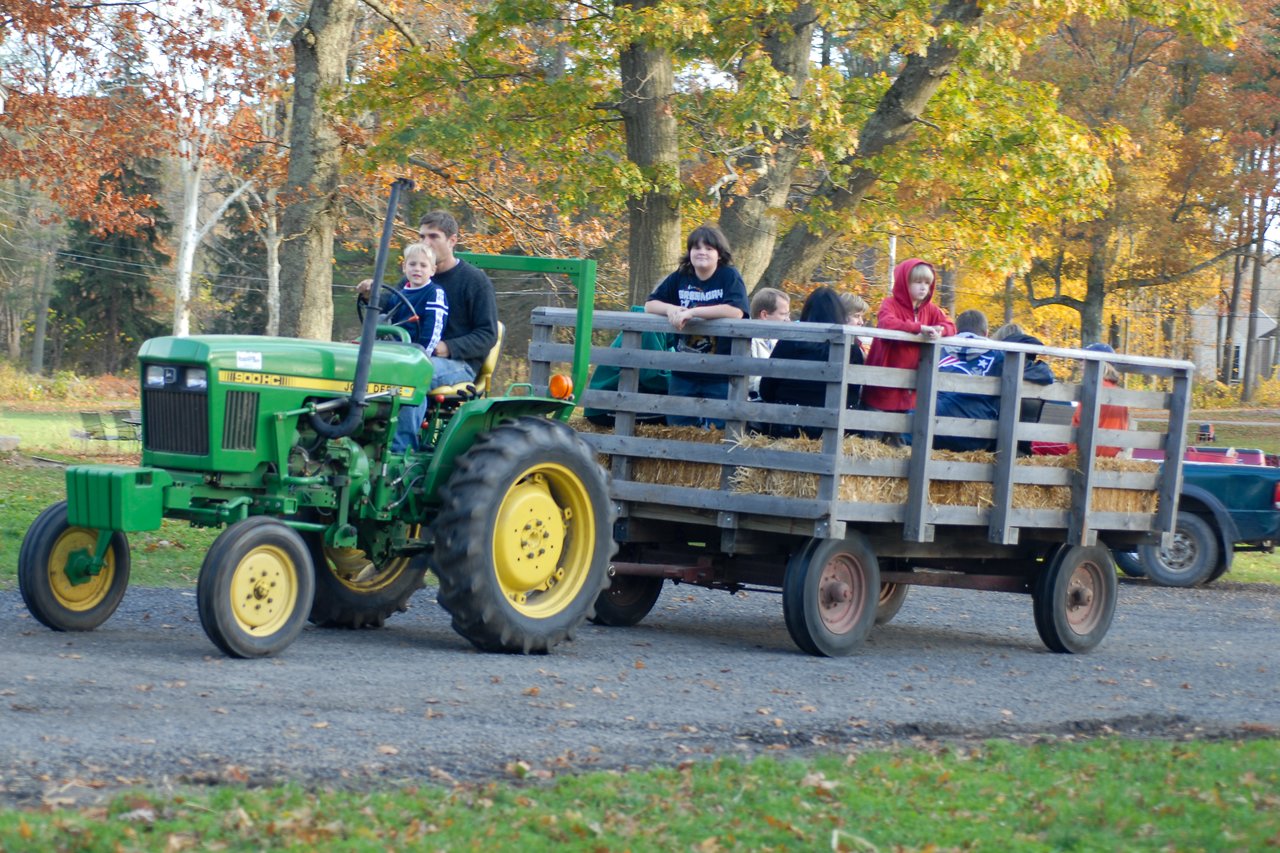 A green tractor pulls a hay wagon with people sitting on straw bales during a hayride in autumn.