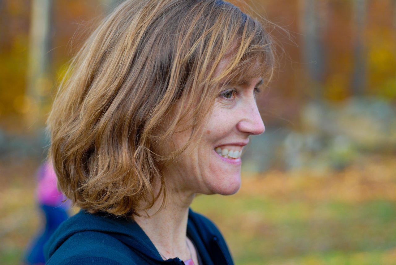A woman with shoulder-length hair smiles while looking to the side at an outdoor gathering in autumn.
