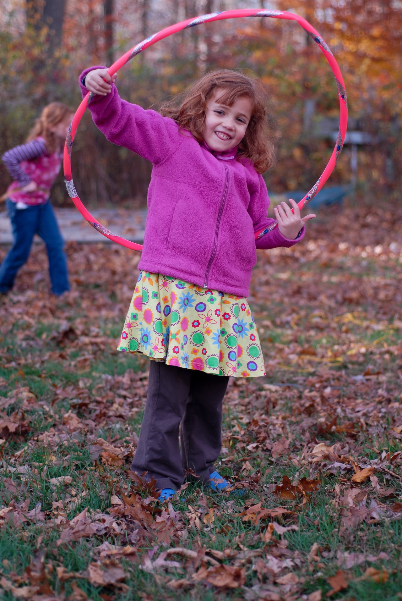 A smiling child in a pink jacket holds a hula hoop outdoors, surrounded by autumn leaves.